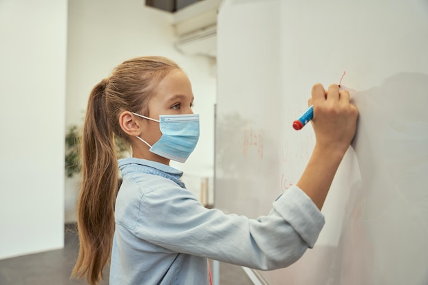 Ritratto di una scolaretta intelligente che indossa una maschera protettiva durante la scrittura della pandemia di coronavirus