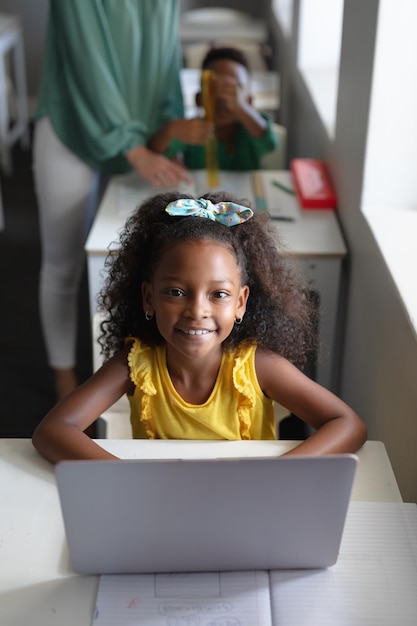 Ritratto di una scuola elementare afroamericana sorridente che usa un portatile alla scrivania in classe