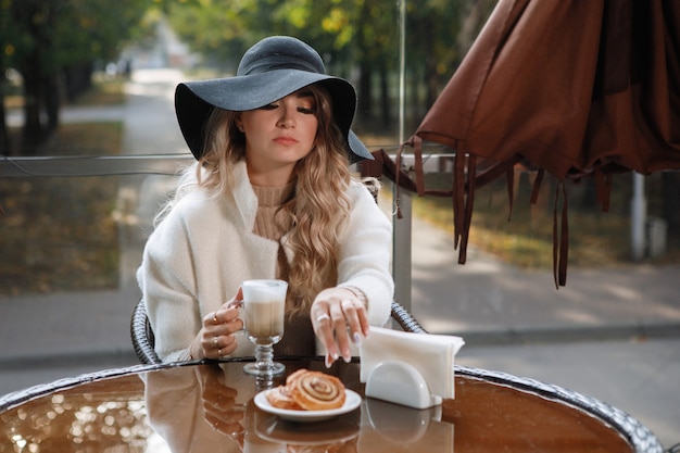Ritratto di una signora bionda con un cappello nero a un tavolo in un caffè che mangia una torta, capelli lunghi, un cappotto beige.