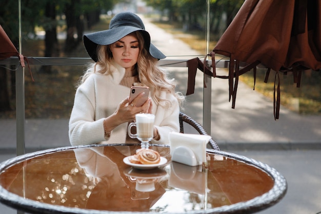 Ritratto di una signora bionda con un cappello nero a un tavolo in un caffè con uno smartphone, capelli lunghi, un cappotto beige. Chiacchierando nei social network, leggendo le notizie.