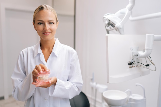 Ritratto di una sorridente ortodontista bionda in cappotto bianco che tiene in mano mascelle umane modello di layout con apparecchi dentali metallici in una moderna clinica dentale Concetto di ortodonzia stomatologica