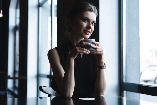 Ritratto di una splendida donna che beve tè o caffè e guarda con un sorriso fuori dalla finestra della caffetteria mentre si gode il suo tempo libero, bel pranzo di una donna d'affari in un caffè moderno durante la sua pausa di lavoro.