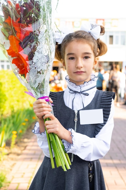 Ritratto di una studentessa della scuola di 1a elementare con fiocchi bianchi e fiori in un'uniforme con badge mock up Ritorno a scuola 1 settembre Prima elementare Classe elementare di istruzione primaria