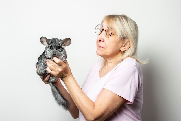 Ritratto di una vecchia amichevole donna in una maglietta e occhiali con un cincillà in mano contro un muro di luce isolato. Volto emotivo. Concetto di animali domestici