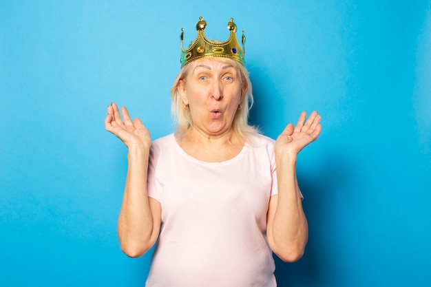 Ritratto di una vecchia donna amichevole con una faccia sorpresa in una maglietta casual con una corona in testa su una parete blu isolata. Volto emotivo