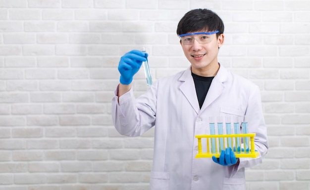 Ritratto di uno scienziato maschio professionista asiatico in camice bianco guanti di gomma e occhiali di sicurezza stanno guardando sorridente la fotocamera che tiene mostrando il rack per provette campione nelle mani su sfondo muro di mattoni.