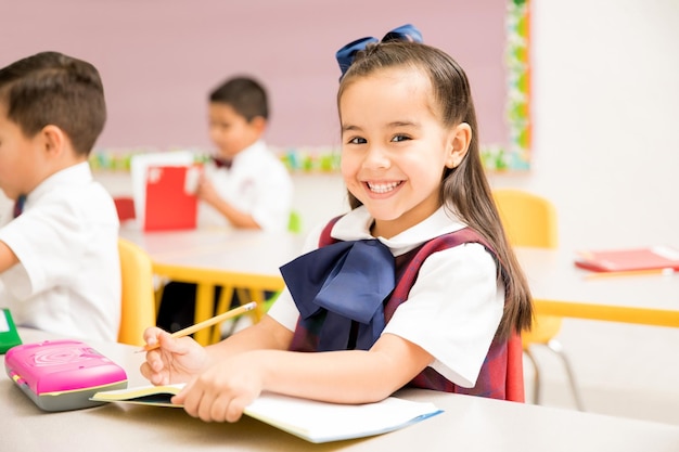 Ritratto di uno splendido studente in età prescolare latino in uniforme che fa un compito di scrittura in un'aula e sorridente