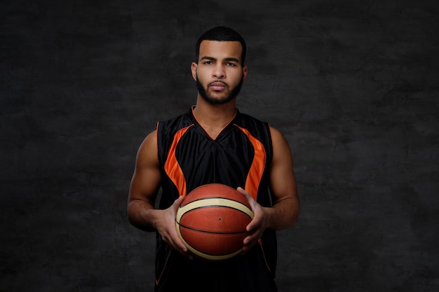 Ritratto di uno sportivo afroamericano. Giocatore di basket in abbigliamento sportivo con una palla su sfondo scuro.