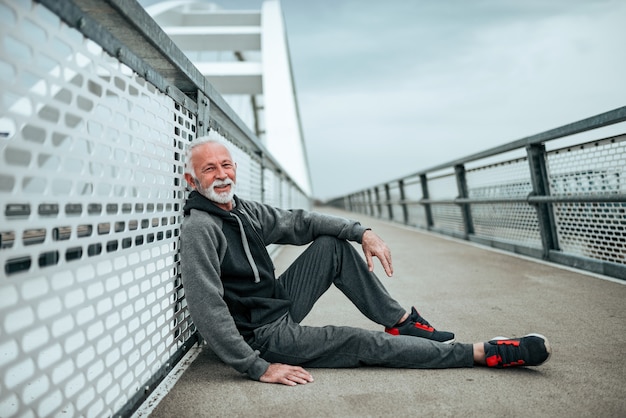 Ritratto di uno sportivo anziano rilassante dopo una corsa. Seduto sul ponte della città, guardando la fotocamera.