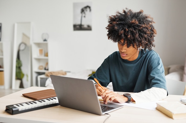 Ritratto di uno studente afroamericano maschio che utilizza il computer portatile mentre fa i compiti nello spazio della copia del dormitorio del college