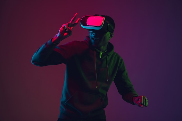 Ritratto di uomo afro-americano in cuffia VR isolato su sfondo sfumato studio in luce al neon. Bellissimo modello maschile. Concetto di emozioni umane, espressione facciale, vendite, pubblicità, inclusione, tecnologia.