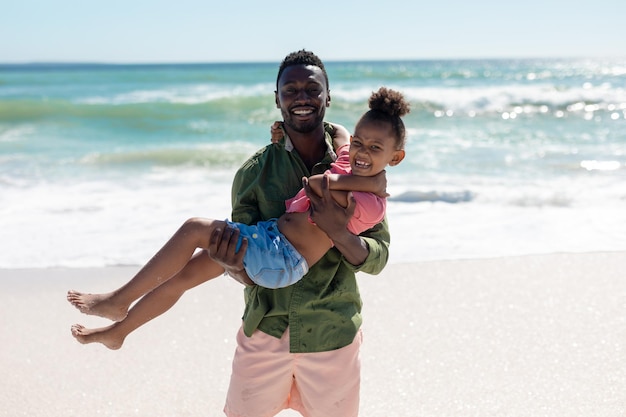 Ritratto di uomo afroamericano che porta la figlia in spiaggia contro il mare in giornata di sole