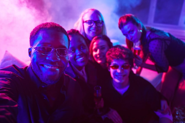 Ritratto di uomo afroamericano che si fa selfie con un gruppo eterogeneo di amici alla festa con luci al neon