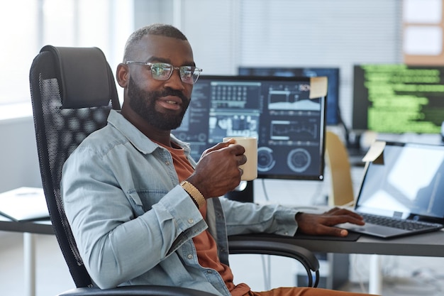 Ritratto di uomo afroamericano come ingegnere del software che guarda la fotocamera mentre posa sul posto di lavoro wi