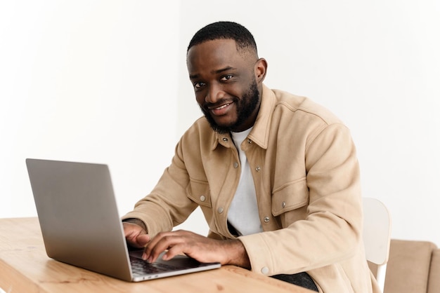 Ritratto di uomo afroamericano sorridente che guarda la fotocamera mentre è seduto al tavolo in ufficio a casa e lavora al computer portatile Felice freelance nero in posa sul posto di lavoro a casa