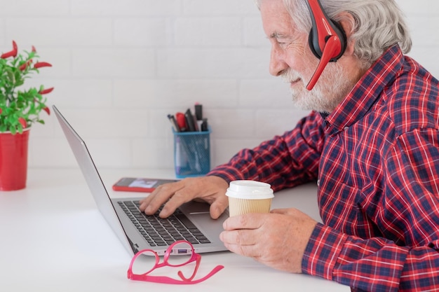 Ritratto di uomo anziano che naviga nei contenuti dei social media sul laptop con in mano una tazza di caffè Vecchio uomo anziano barbuto che trascorre il suo tempo lavorando al computer a casa usando le cuffie