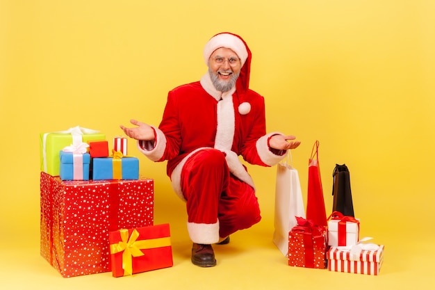 Ritratto di uomo anziano con barba grigia in costume di babbo natale che mostra molte scatole e borse della spesa presenti, pronto a congratularsi con il nuovo anno. Colpo dello studio dell'interno isolato su fondo giallo