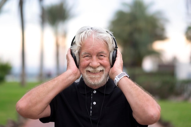 Ritratto di uomo anziano dai capelli bianchi felice con la barba che indossa le cuffie per ascoltare musica sorridente