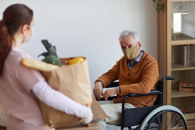 Ritratto di uomo anziano in sedia a rotelle che indossa una maschera mentre accetta la consegna di cibo, il concetto di aiuto e assistenza, spazio di copia