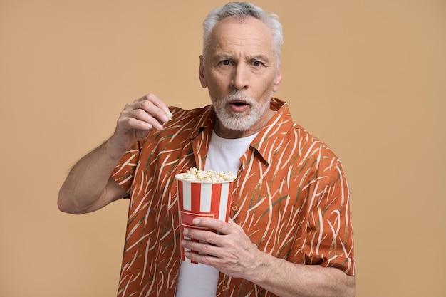 Ritratto di uomo anziano stupito che mangia popcorn guardando video divertendosi su sfondo beige