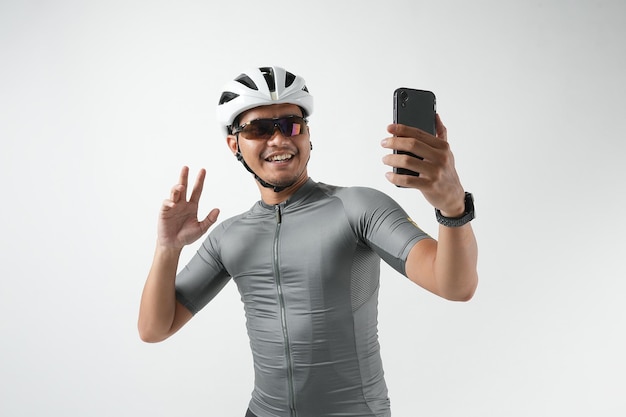 Ritratto di uomo asiatico che indossa t-shirt e casco da bici bianco, effettua videochiamate e saluta il telefono