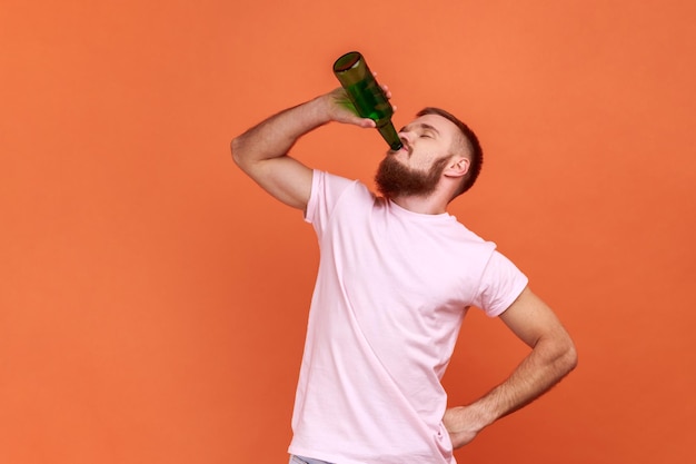 Ritratto di uomo barbuto in piedi con una bottiglia di birra ubriaco in piedi con gli occhi chiusi e bere alcolici indossando maglietta rosa Indoor studio girato isolato su sfondo arancione