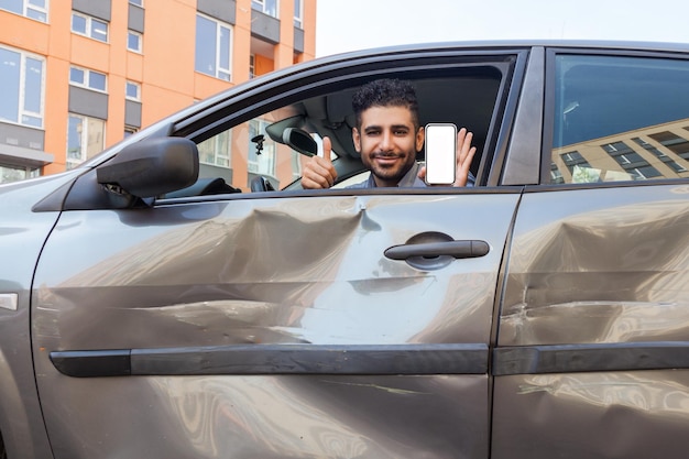 Ritratto di uomo barbuto in un'auto schiantata che guarda la fotocamera e sorride, mostrando il pollice in su e smartphone con schermo vuoto, servizi di ispezione assicurativa pubblicitaria dopo un incidente stradale. Tiro all'aperto.