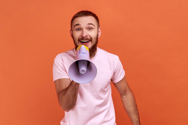 Ritratto di uomo barbuto ottimista che urla nel megafono che annuncia informazioni importanti con una fotocamera positiva che indossa una maglietta rosa Indoor studio girato isolato su sfondo arancione