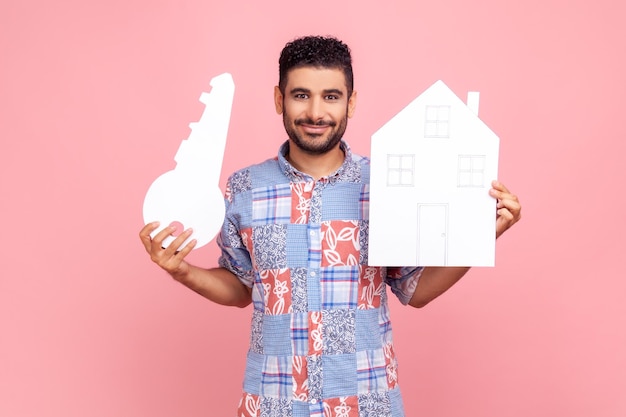 Ritratto di uomo barbuto soddisfatto in camicia casual blu che tiene casa di carta e mutuo per l'acquisto di immobili chiave di grandi dimensioni Studio indoor girato isolato su sfondo rosa