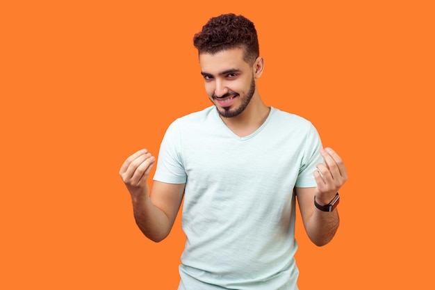 Ritratto di uomo castano bello positivo con la barba in t-shirt bianca che guarda giocoso alla macchina fotografica e mostra gesto italiano o segno di denaro con le mani. tiro in studio indoor isolato su sfondo arancione