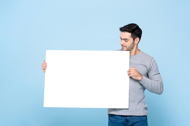 Ritratto di uomo caucasico bello che tiene e guarda il cartello bianco in uno sfondo blu studio isolato