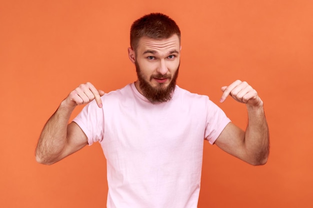 Ritratto di uomo che punta le dita verso il basso prestando attenzione all'area pubblicitaria che mostra lo spazio libero guardando la fotocamera con indosso una maglietta rosa Indoor studio girato isolato su sfondo arancione