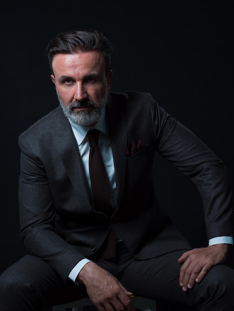 Ritratto di uomo d'affari adulto che indossa un abito alla moda e seduto in uno studio moderno su una sedia elegante sullo sfondo nero. Modello orizzontale. Foto di alta qualità