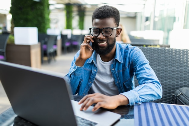 Ritratto di uomo d'affari africano felice utilizzando il telefono mentre si lavora al computer portatile in un ristorante