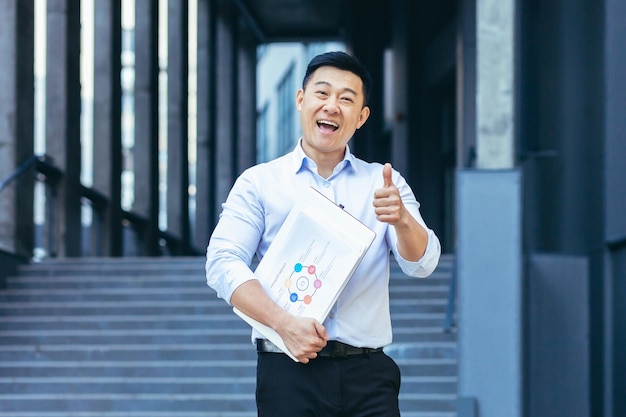 Ritratto di uomo d'affari asiatico broker uomo fuori ufficio tenendo laptop e documenti con grafici guardando la fotocamera sorridente tenendo i pollici in su