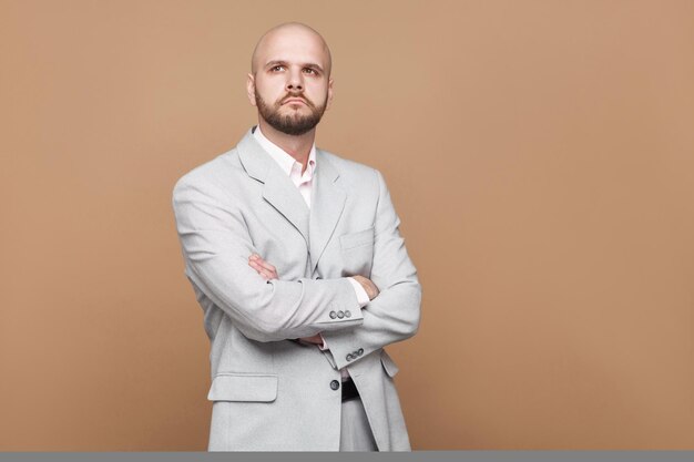 Ritratto di uomo d'affari barbuto calvo di mezza età bello confuso nel classico abito grigio chiaro in piedi e pensando con le mani incrociate. studio indoor girato, isolato su sfondo marrone chiaro.