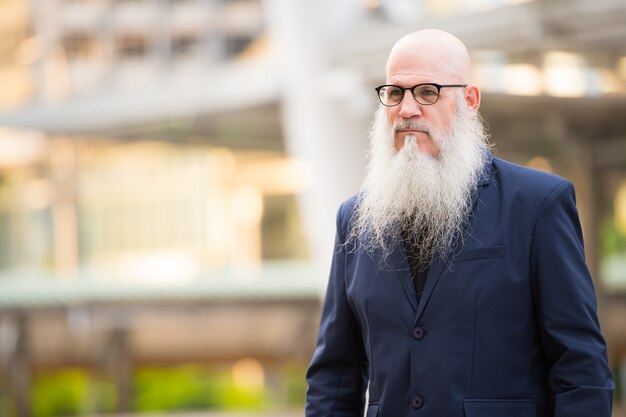 Ritratto di uomo d'affari calvo maturo con barba lunga che indossa occhiali da vista nelle strade della città all'aperto