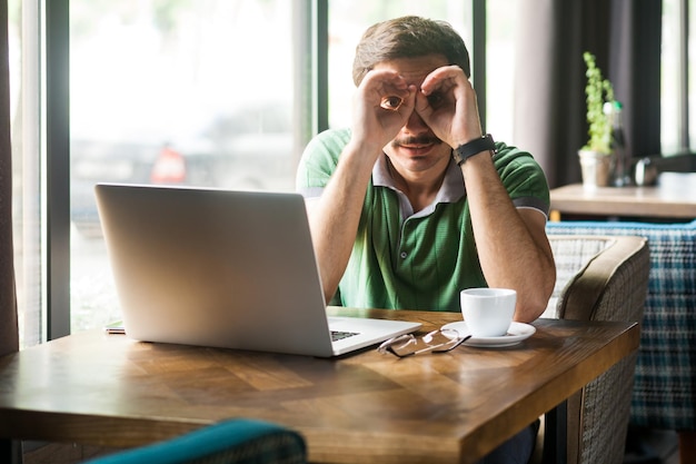 Ritratto di uomo d'affari che indossa una maglietta verde, lavora online su laptop, guarda attraverso le dita a forma di binocolo ed esprime positivo. Ripresa in interni vicino alla grande finestra, sfondo del caffè.