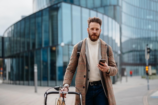 Ritratto di uomo d'affari hipster con bici utilizzando il cellulare e guardando la fotocamera Posizione aziendale