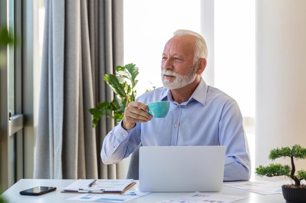 Ritratto di uomo d'affari senior sorridente che tiene tazza di caffè guardando attraverso la finestra vicino spazio libero