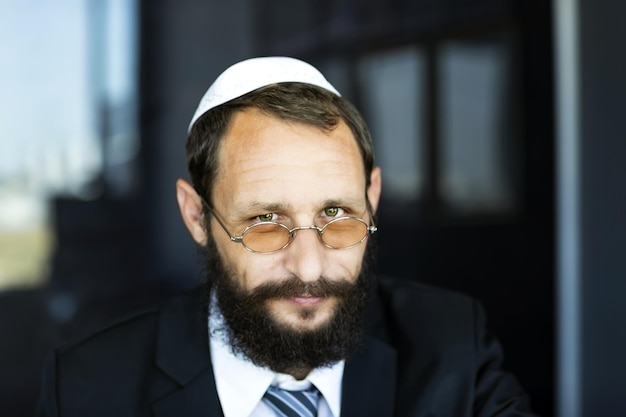 Ritratto di uomo ebreo barbuto affascinante in cappello bianco Yarmulke Kippah Espressione emotiva Colpo alla testa uomo ebreo barbuto serio che guarda la telecamera Messa a fuoco selettiva sugli occhi Concetto di occhiali