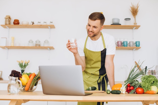 Ritratto di uomo felice a casa uomo che cucina le verdure e mostra un bicchiere d'acqua che affetta le verdure utilizzando il laptop per la formazione di cucina online