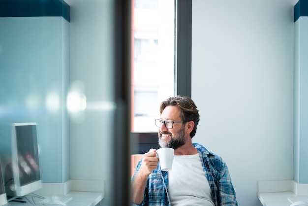 Ritratto di uomo felice che guarda fuori dalla finestra e sorride bevendo un caffè a casa o in ufficio - maschio caucasico adulto con barba e occhiali nell'attività di pausa lavorativa da solo - persone felici con gli occhiali