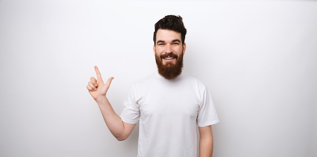 Ritratto di uomo felice con la barba in maglietta bianca che punta lontano su sfondo bianco