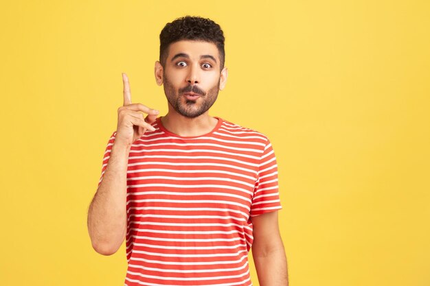 Ritratto di uomo felice eccitato con la barba in camicia a righe che punta il dito verso l'alto scopri una soluzione o un'idea ottieni una risposta alla domanda eureka Indoor studio shot isolato su sfondo giallo