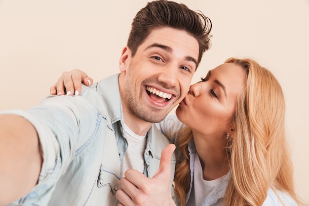 Ritratto di uomo felice prendendo selfie foto e gesticolando pollice in alto mentre adorabile donna che bacia la sua guancia