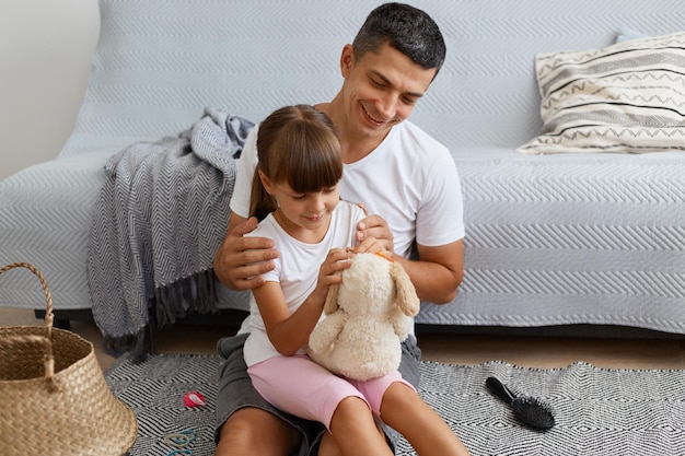 Ritratto di uomo felice sorridente che indossa maglietta bianca e jeans in stile casual corto seduto sul pavimento con la figlia piccolo bambino che tiene insieme la famiglia di peluche trascorrendo il fine settimana