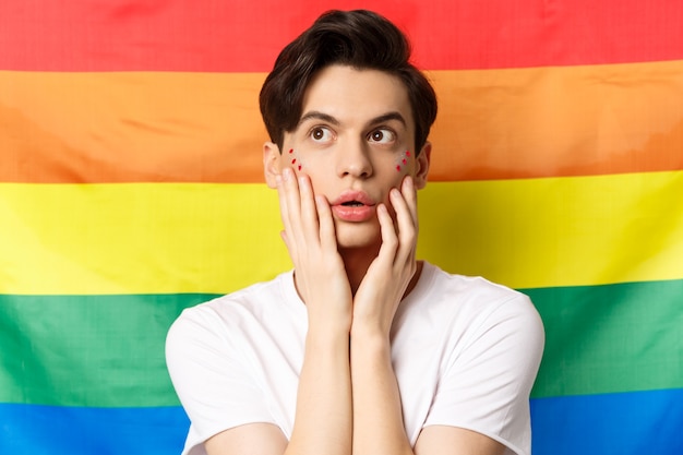 Ritratto di uomo gay sensuale e bello con trucco glitter sul viso, guardando l'angolo in alto a sinistra eccitato, in piedi contro la bandiera arcobaleno lgbtq.