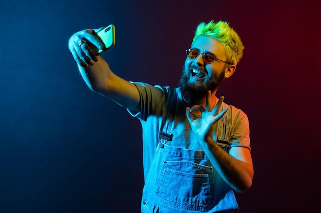 Ritratto di uomo hipster felice con la barba che fa selfie tramite smartphone, mostrando segno di approvazione ok alla fotocamera dello smartphone, live streaming, colloqui con i follower. Luce al neon colorata, ripresa in studio al coperto.