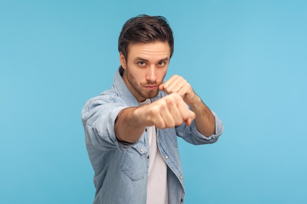 Ritratto di uomo in camicia di jeans da lavoratore in piedi con gesto di boxe, pugni alla telecamera, provocando lotta o pronto all'autodifesa, concetto di lotta. tiro in studio indoor isolato su sfondo blu